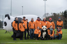 Komm auch du in unser Freiwilligenteam! Zusammen Gutes tun ist sinnvoll und macht Spass! Zwölf freiwillige Mitarbeitende posieren vor dem aufgeblasenen Pandabär, anlässlich eines WWF-Laufes.