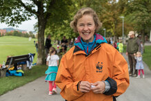 Ein Freiwilligeneinsatz an einem WWF-Lauf ist sinnvoll und macht garantiert Freude! Eine freiwillige Helferin motiviert die Kinder an einem WWF-Lauf. Sie trägt eine orange WWF-Jacke und lächelt in die Kamera, im Hintergrund rennen die Kinder.